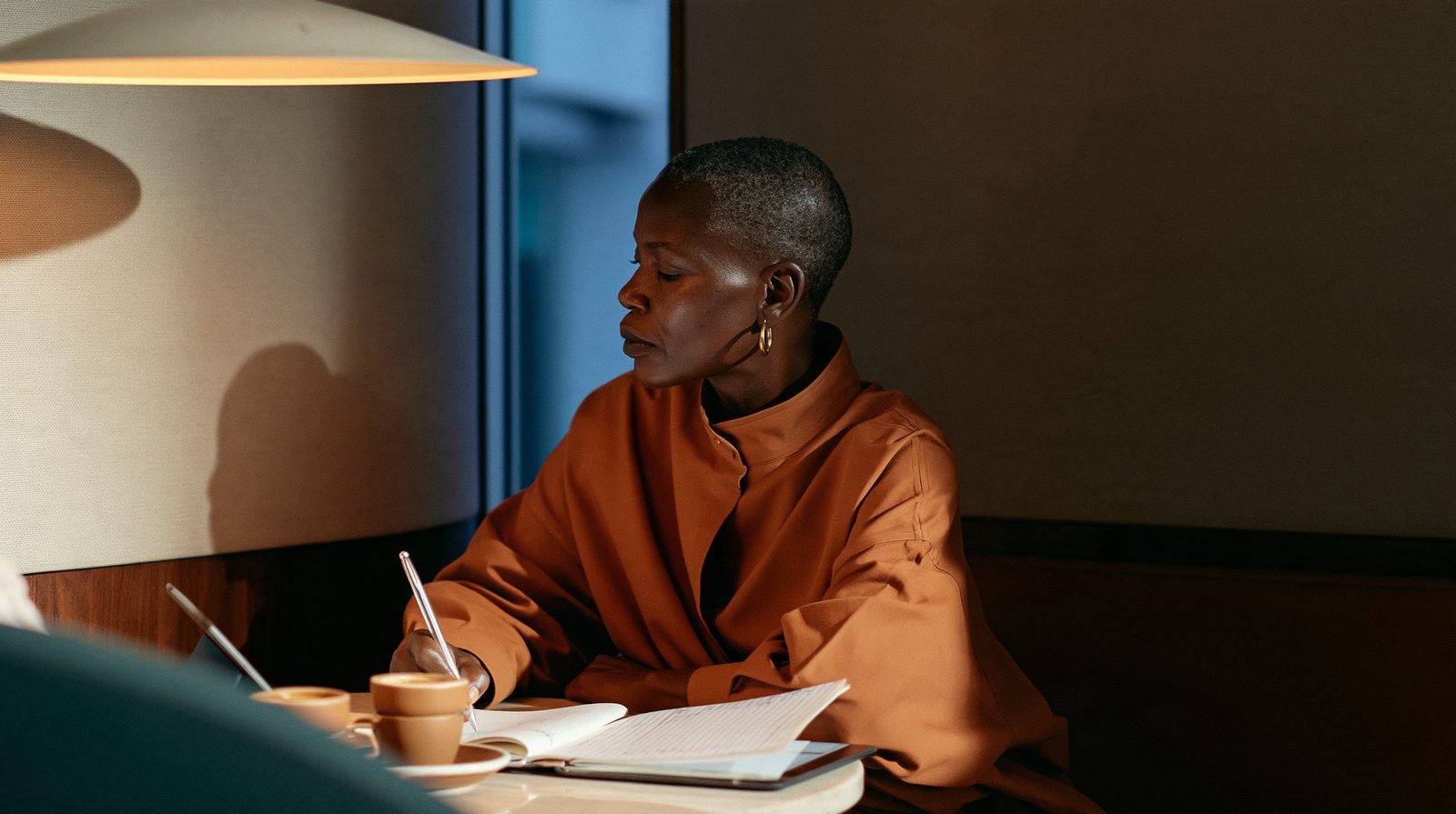 A woman writes in a ledger at a small round café table, espresso cups beside her tablet, cool dusk blue at the window.