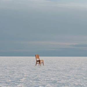 A single wooden chair stands alone on a desolate, cracked salt flat under an overcast, pale gray sky.