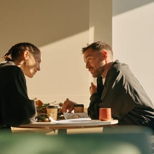 A florist with braided hair and a café owner lean across a small round table, loose stems and a printed plan between their coffee cups.