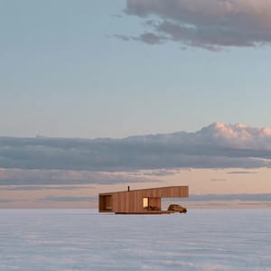A minimalist cabin stands perfectly centered on a white salt flat, split by a stark horizon bisection under a pale sky.