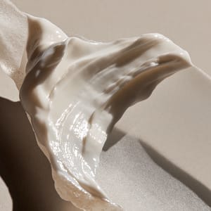 Macro shot of a thick white lotion swatch smeared across frosted glass, highlighting its rich and glossy hydrating texture.