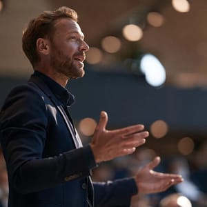 A motivational man in a sharp suit gestures passionately on a bright stage, inspiring an audience with a confident smile.