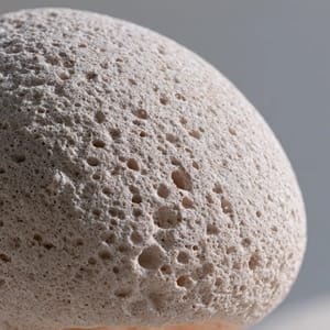 A macro close-up of a sphere carved from pumice, its rough porous surface covered in tiny craters, illuminated by soft diffuse light.