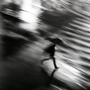 A lone pedestrian crosses a dark night street, rendered in rough blurry monochrome with heavy film grain.