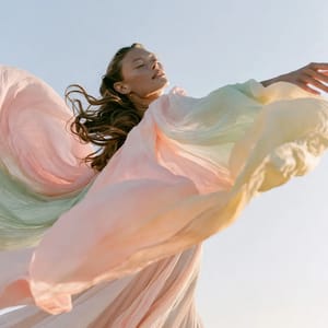 A woman in a flowing dress stands against a pastel backdrop, the fabric blending pale pink, mint green, and butter yellow in a Spring palette.