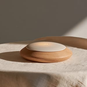 A close-up of a minimalist handheld smart device featuring tactile technology, combining a smooth light oak chassis with a soft matte e-ink display resting on a linen surface.
