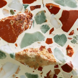 A polished slab of terrazzo aggregate with irregular chips of emerald green, ochre, and white marble embedded in a cream-colored cement base.