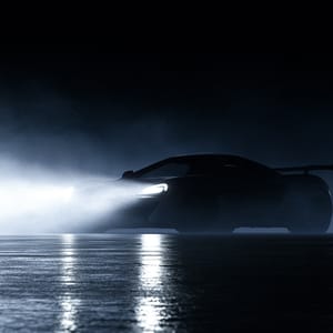 A sports car on wet asphalt at night cuts through misty air with a bright volumetric headlight beam.