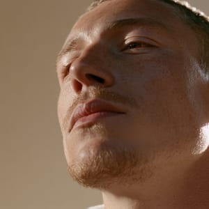 A close-up of a man's face in quiet contemplation, bathed in wrapped soft light against a warm beige background.