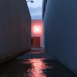 One-point perspective of a quiet, empty concrete alleyway at dusk. Smooth concrete walls on both sides converge toward a single glowing neon sign at the far end, casting a warm red reflection on the wet pavement. Clean, dramatic linear composition. --ar 16:9 --style raw