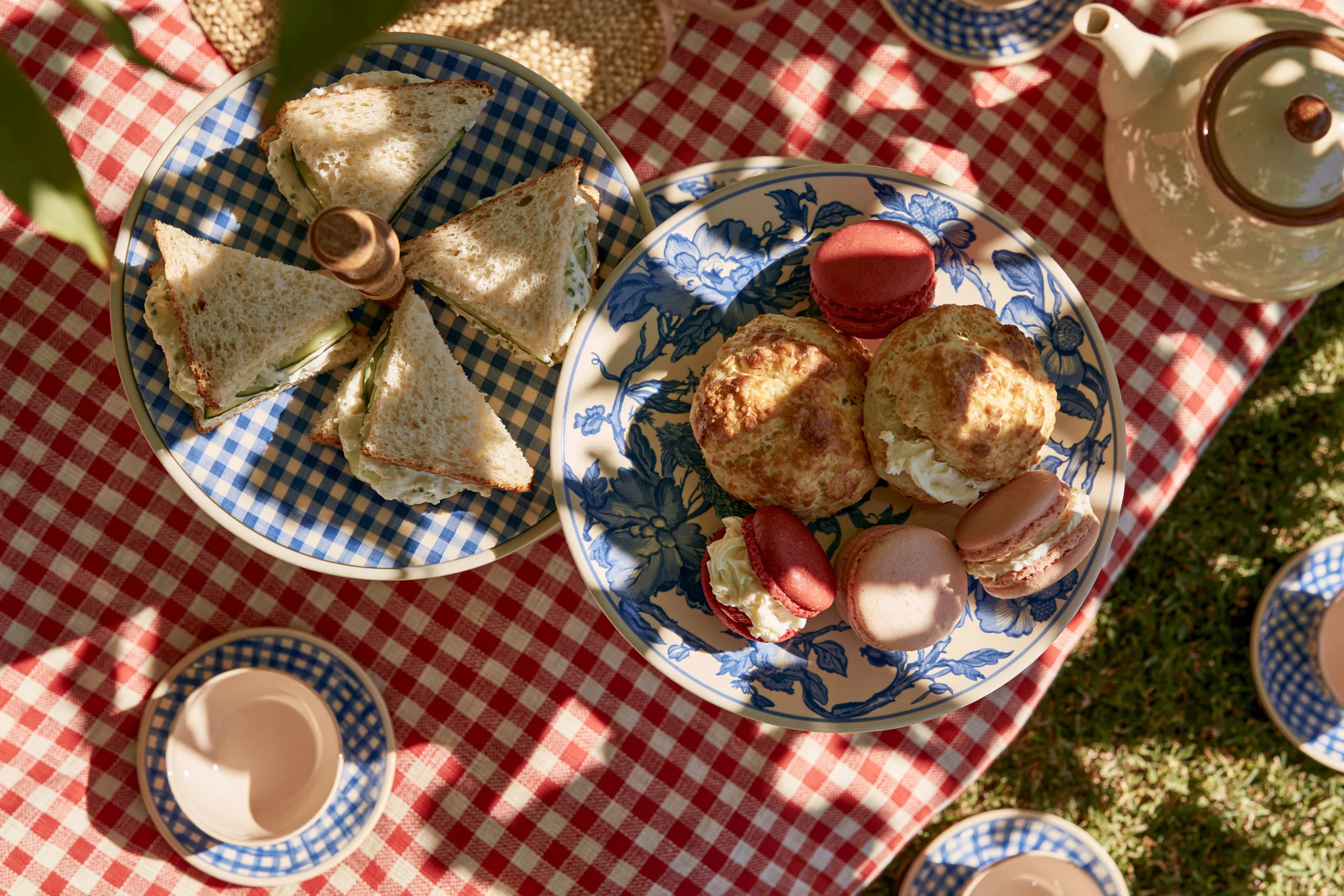 An overhead shot of a three-tiered afternoon tea stand filled with pastries on a checkered picnic blanket in sunny grass.