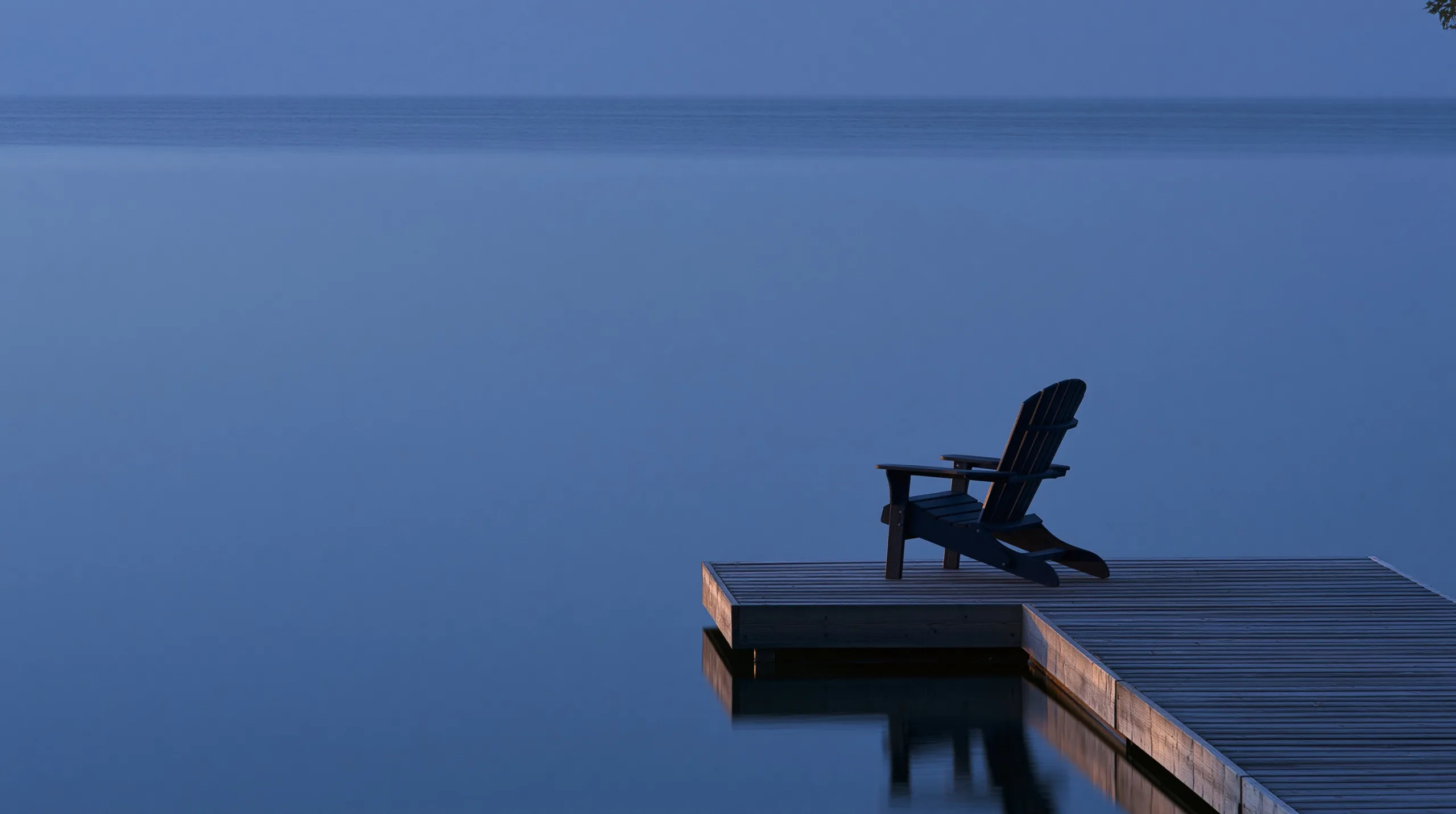 A solitary wooden chair sits on a quiet lakeside pier, enveloped in the soft, cool tones of the blue hour.