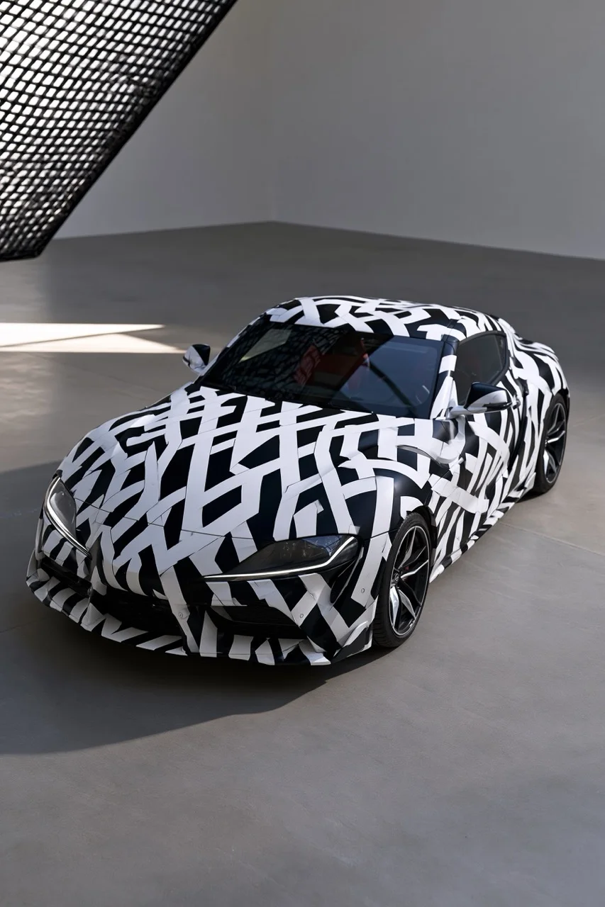 A sleek aerodynamic sports car painted with dazzle camouflage, featuring sharp intersecting black and white geometric shapes disrupting its flowing curves in a smooth gray concrete studio.