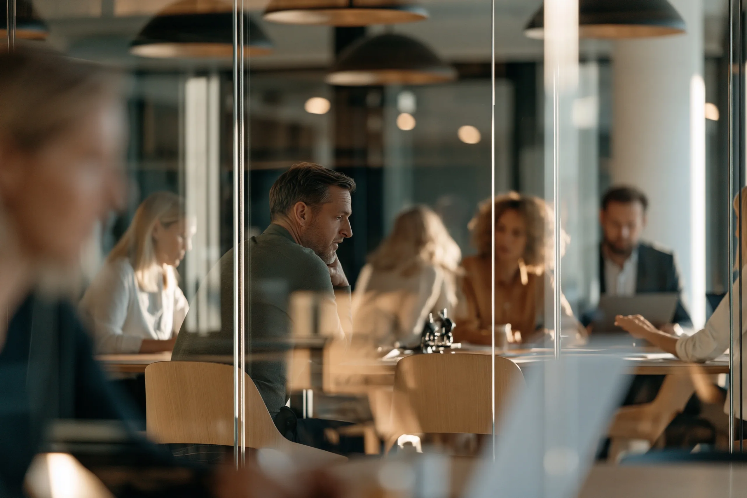 A modern collaborative meeting room viewed through a frameless glass partition with subtle light reflections showing professional teamwork in action.