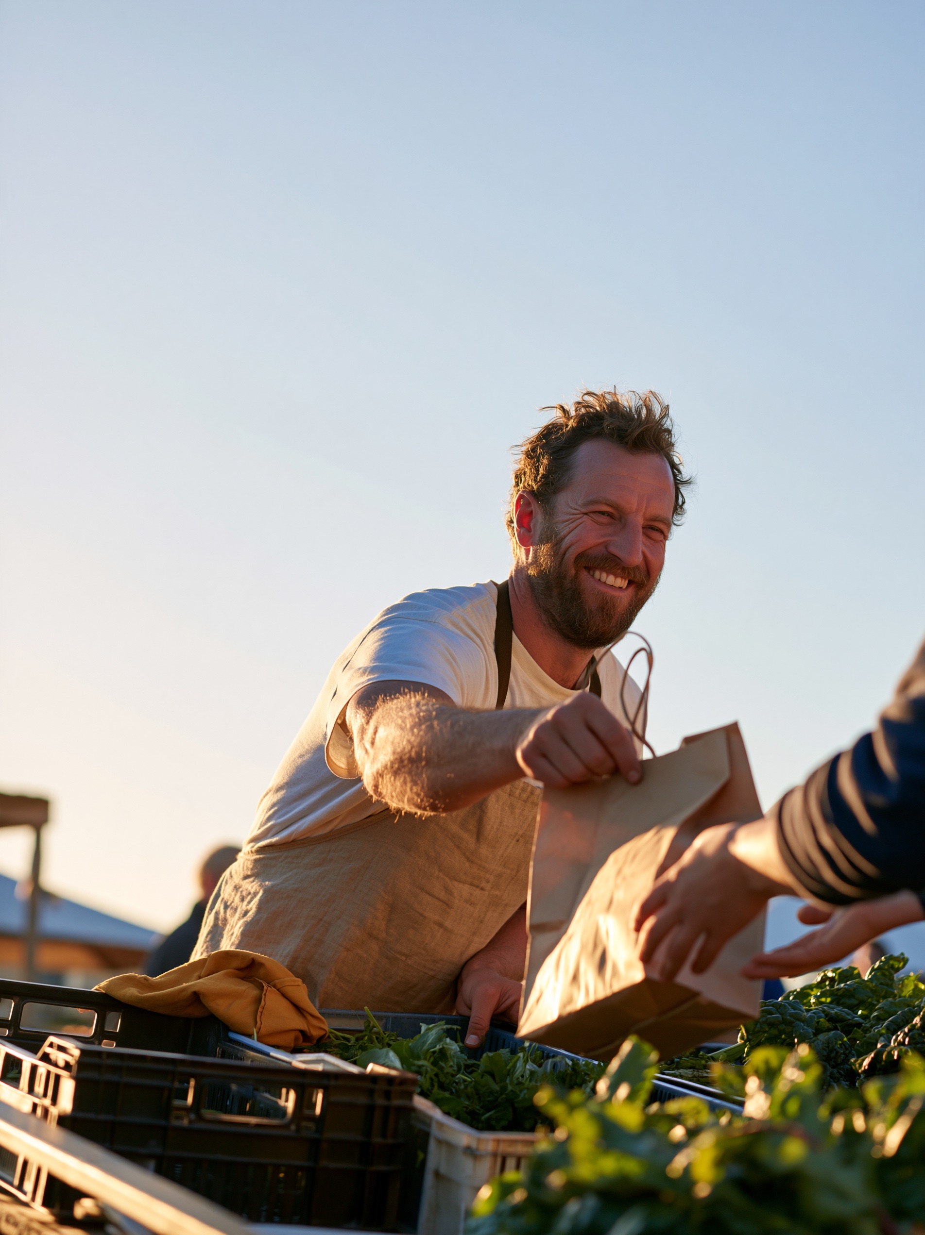 A grower in a linen apron passes a kraft paper bag of leafy greens to a customer's hands across a market table.