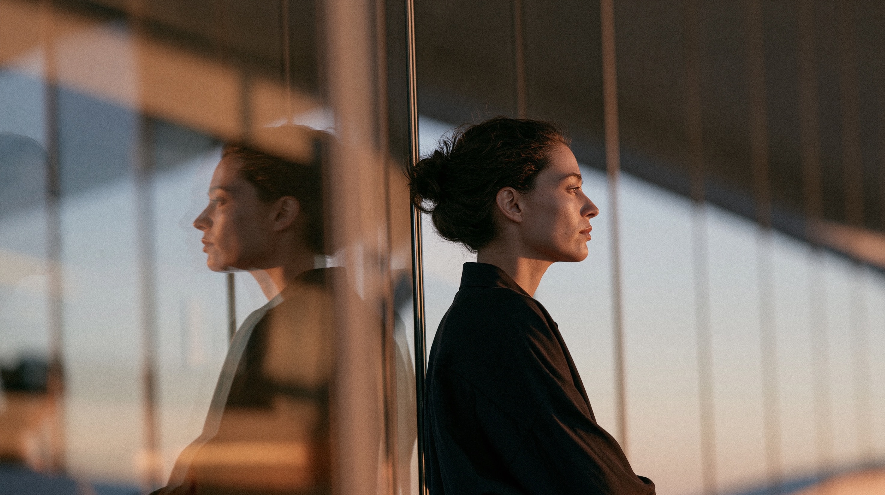 A soft profile of a woman seen through multiple window panes, creating a complex layered glass reflection in muted dusk light.