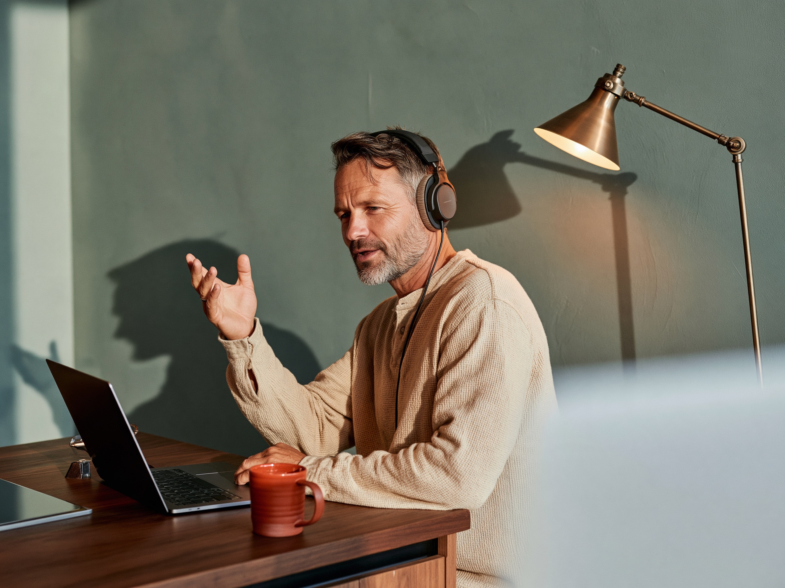A bearded man in his forties gestures mid-sentence at a kitchen island, laptop and headphones on, lit by a warm lamp.
