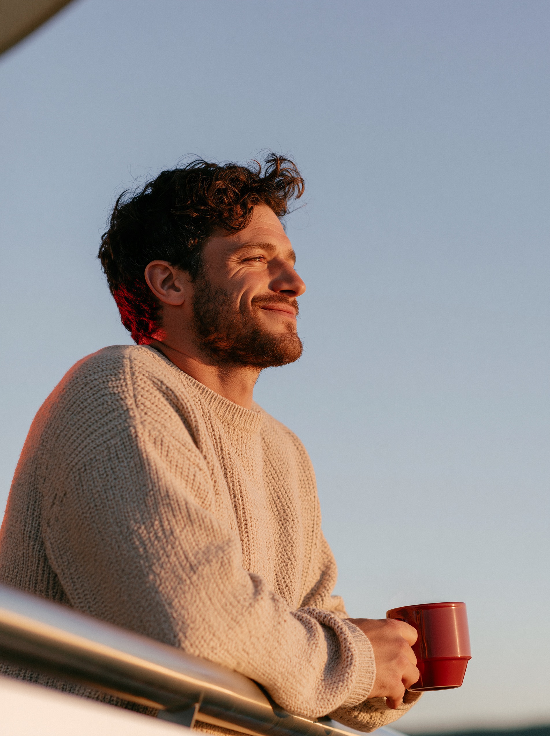 A man in an oatmeal sweater holds a steaming mug by a steel railing against a soft golden sunset sky.