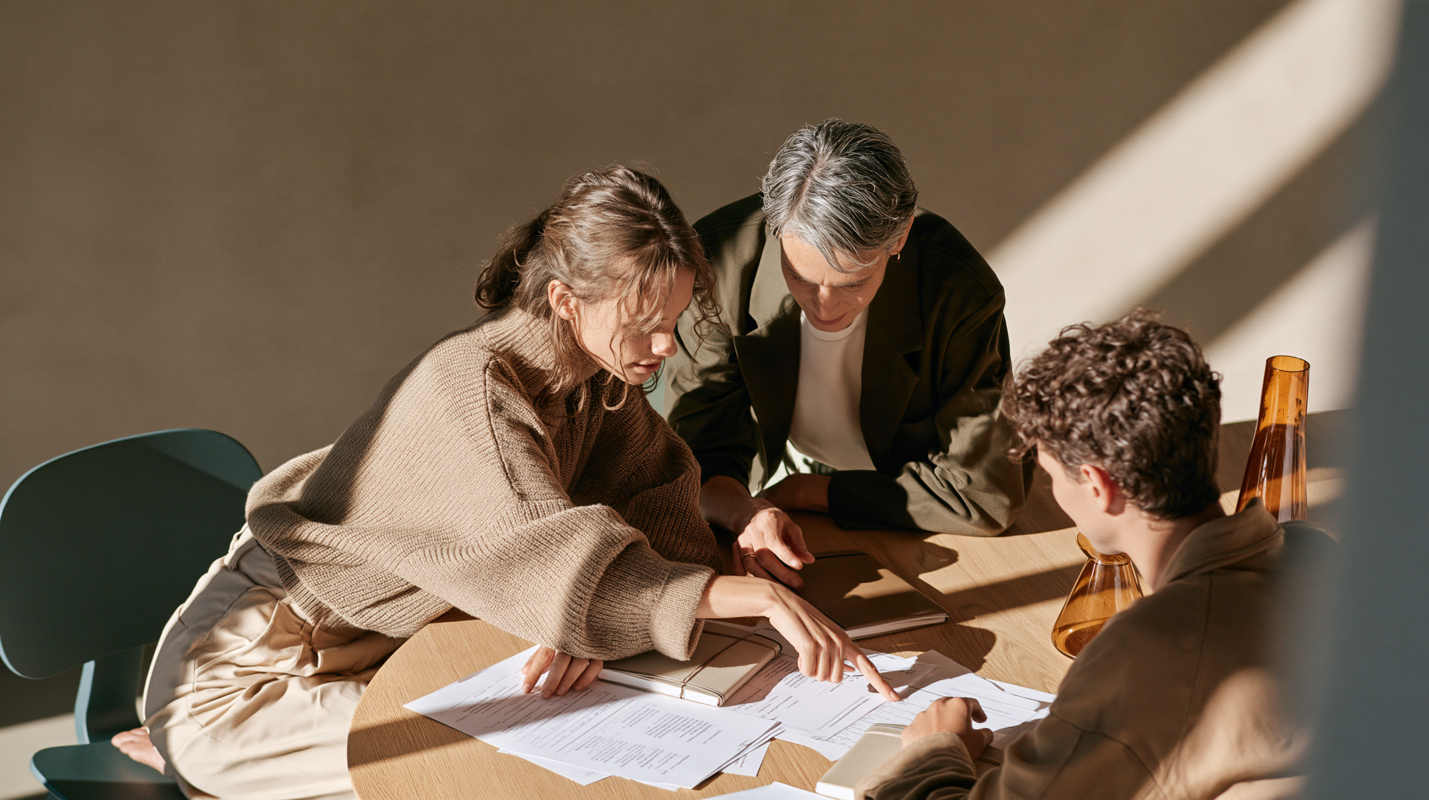 Three colleagues lean over a round oak table with printed agendas, one pointing at a line as soft light falls from the right.