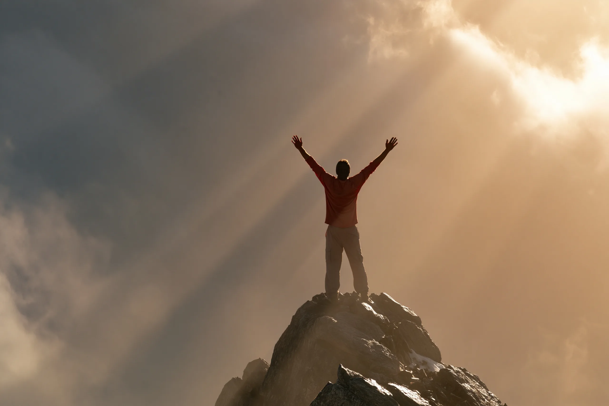 A triumphant figure stands at the peak of a rugged summit, arms raised in silhouette against golden sunbeams piercing through dispersing clouds, the upward sweep of the pose echoing the mountain's upw