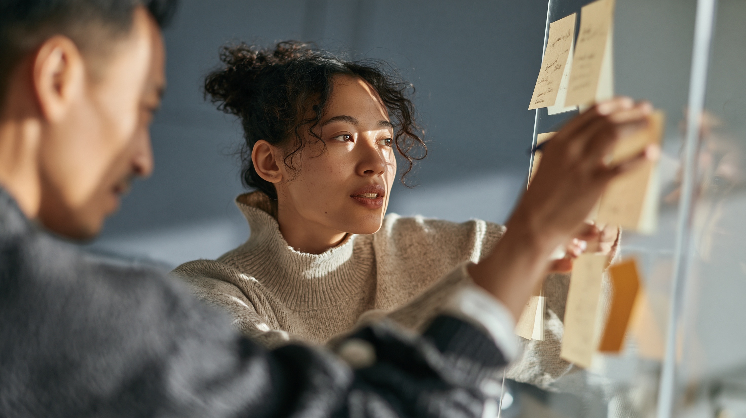 Over the shoulder view of two colleagues arranging notes on an office glass wall with soft lighting and open wall space.