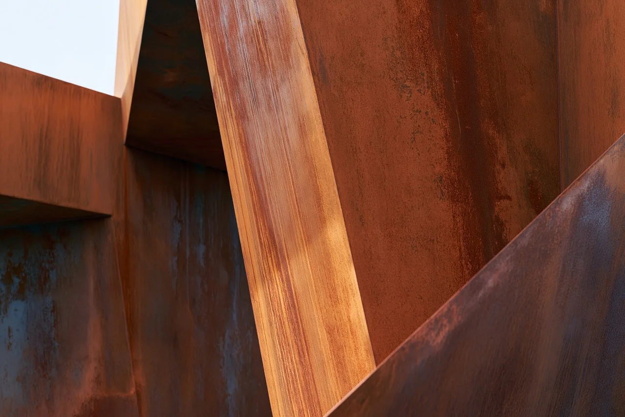 A minimalist architectural pavilion constructed from weathering steel plates set against a moody, overcast coastal landscape.