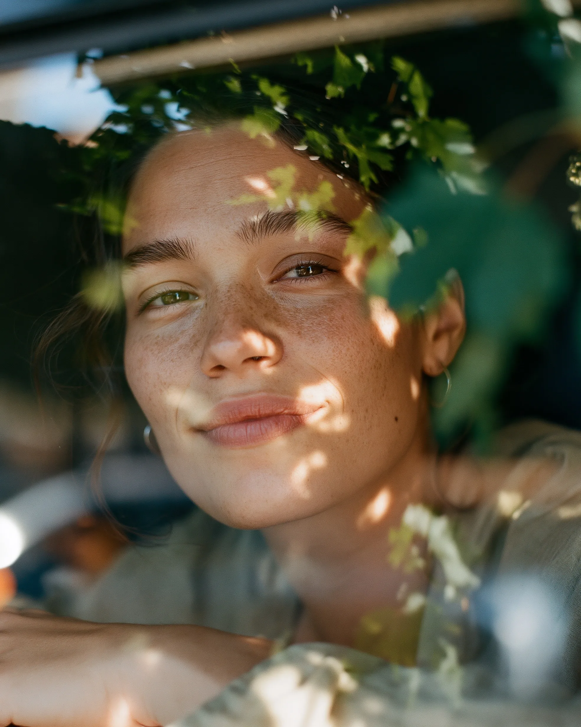 A quiet portrait of a woman seen through a window reflection of soft green leaves and gentle afternoon light.