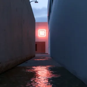 One-point perspective of a quiet, empty concrete alleyway at dusk. Smooth concrete walls on both sides converge toward a single glowing neon sign at the far end, casting a warm red reflection on the wet pavement. Clean, dramatic linear composition. --ar 16:9 --style raw